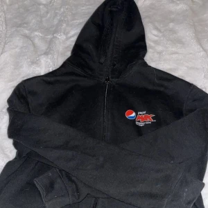Svart Pepsi Max hoodie med dragkedja - Svart hoodie med Pepsi Max-logga på bröstet. Tröjan har huva med snörning, långa ärmar och dragkedja framtill. Tillverkad i mjuk bomull och polyesterblandning, perfekt för chill dagar. Klassisk passform och ribbade muddar vid ärmslut och nederkant. 