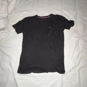 Svart t-shirt från Tommy Hilfiger - Svart t-shirt från Tommy Hilfiger med liten logga på bröstet. Klassisk rund hals och korta ärmar. Skön passform och tillverkad i mjuk bomull. Perfekt för en clean och enkel stil.