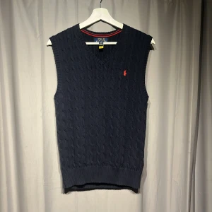 Mörkblå stickad väst från Polo Ralph Lauren - Snygg mörkblå stickad väst från Polo Ralph Lauren med klassisk kabelstickning och röd broderad logga på bröstet. V-ringad hals och ribbad kant nedtill ger en clean look. Perfekt att slänga över en skjorta. Nypris 1000kr