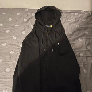 Svart hoodie från Polo Ralph Lauren - Svart hoodie från Polo Ralph Lauren med klassisk vit broderad logga på bröstet. Tröjan har huva med snörning, dragkedja framtill och är tillverkad i mjuk bomull. Perfekt för dig som gillar stilren och enkel design.