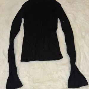 Svart ribbad polotröja från Stradivarius - Snygg svart polotröja från Stradivarius i ribbstickad design. Tröjan har långärmade, utsvängda ärmar och en tight passform som sitter riktigt snyggt. Perfekt för dig som gillar stilrena och trendiga plagg med lite edge.