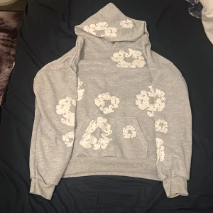 Grå hoodie från Denim Tears med mönster - Grå hoodie från Denim Tears med vita, abstrakta mönster tryckta över hela tröjan. Tröjan har huva, magficka och ribbade muddar vid ärmslut och nederkant. Perfekt för dig som gillar streetwear och unika prints.