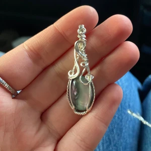 Silverberlock med ”norrskenssten”  - Unik berlock med en oval, gråskimrande sten infattad i intrikat virad silvertråd. Hängsmycket har snygga snirkliga detaljer och en elegant, bohemisk vibe. Perfekt för dig som gillar statement-smycken med konstnärlig känsla.