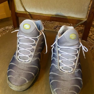 Nike Air Max Plus grå och gul sneakers - Nike Air Max Plus sneakers i grått och gult med vita snören och svarta detaljer. Ovandelen har ett vågigt mönster och meshmaterial, med synlig Air Max-dämpning i sulan. Snyggt TN-märke på hälen och gul Swoosh på sidan. Perfekt för dig som gillar retrostil och streetwear.