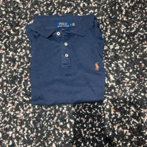 Blå Polo Ralph Lauren piké slim fit - Snygg blå pikétröja från Polo Ralph Lauren i slim fit-modell. Klassisk krage, tre knappar framtill och den ikoniska broderade loggan på bröstet. Kortärmad och tillverkad i mjuk bomull som är skön mot huden. Perfekt för en clean och stilren look.