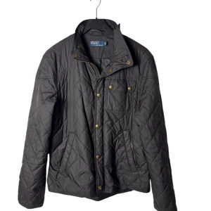 Ralph Lauren quiltad jacka - Svart quiltad jacka från Polo Ralph Lauren med snygga guldfärgade knappar och dragkedja. Jackan har hög krage, fickor med dragkedja och tryckknappar samt ett stilrent rutmönster. Perfekt för dig som gillar klassisk och clean stil.