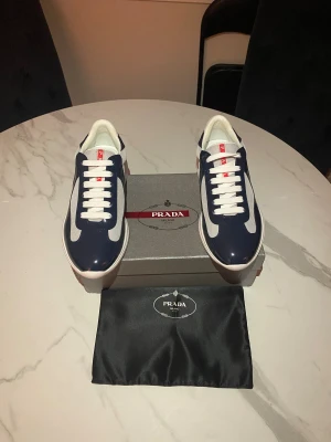 Prada sneakers i marinblå och vit - Snygga Prada sneakers med marinblå och vita paneler, vit sula och röda logodetaljer. Skorna har rund tå, snörning och meshpartier för extra komfort. Kombination av syntet och mesh ger en modern och sportig vibe. Kommer med originalkartong och dustbag.