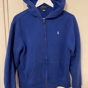 Blå hoodie från Polo Ralph Lauren  - Blå hoodie från Polo Ralph Lauren. Passar i storlek S-M. Klassisk design med dragkedja, huva och kängurufickor. Liten vit logga broderad på bröstet. Perfekt för chill dagar eller när du vill ha en clean streetstyle.