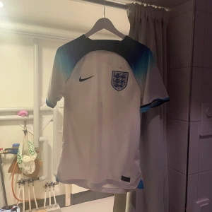 Äkta England Nike fotbollströja S - England fotbollströja från Nike i storlek S. Tröjan är vit med mörkblå och ljusblå detaljer vid axlar och ärmslut, samt England-emblem på bröstet. Tillverkad i polyester med rund hals och korta ärmar. Perfekt för dig som vill representera Three Lions.