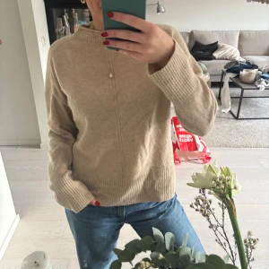 Beige stickad tröja från Esprit - Mysig beige stickad tröja från Esprit med ribbad hög hals och muddar. Tröjan har en rak passform och diskret söm framtill, perfekt till jeans eller kjol. Enkel och stilren design som funkar året runt.