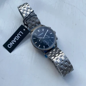 Klcoka - BUTIKSPRIS 2995kr. Stilren klocka från Lugano med svart urtavla och tre mindre subdials. Boetten och armbandet är i blankt silverfärgat rostfritt stål. Klockan har datumvisning och klassisk länkdesign. Perfekt accessoar för dig som gillar en clean och modern look. PRISET KAN DISKUTERAS.