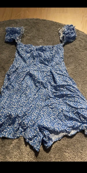 Blåvit blommig kortklänning - Supersöt blå kortklänning med vita blommor och puffiga kortärmar. Klänningen har en figurnära passform upptill och utställd kjol, perfekt för varma dagar. Tunt och lätt material som känns luftigt. Ingen synlig dragkedja eller knappar. Med inbyggda shorts så sitter på plats.
