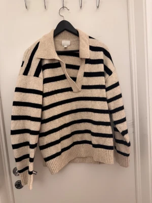 Randig beige och svart stickad tröja  - Snygg stickad polotröja i beige med svarta ränder. Tröjan har en bred krage och djup v-ringning, samt långa ärmar och ribbade muddar vid ärmslut och nederkant. Perfekt för dig som gillar en chill och trendig look. 