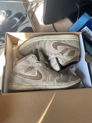 Nike Air Jordan 1 High beige mocka - Nike Air Jordan 1 High sneakers i beige mocka med bruna detaljer och unikt mönstrad swoosh. Skorna har snörning, svart innersula och klassisk Jordan-logga i guld på sidan. Perfekt för dig som gillar retro och streetwear vibes.