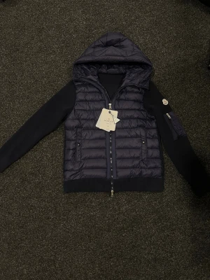 Mörkblå pufferjacka från Moncler - Snygg mörkblå cardigan från Moncler med stickade ärmar och rygg, quiltad framsida och huva. Jackan har dragkedja framtill, fickor med tryckknappar och en extra ficka med dragkedja på ärmen. Moncler-logga på vänster ärm för extra stilpoäng. Aldrig använt på grund av köpt fel storlek😊