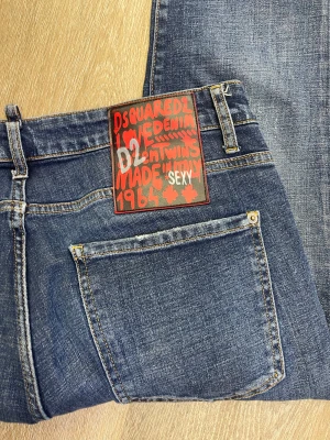 Dsquared2 blå jeans med slitningar - Säljer ett par blå jeans från Dsquared2 med coola slitningar och tvättade detaljer. Klassisk femficksmodell med gul kontrastsöm och röd Dsquared2-tag vid gylfen. Jeansen har en rak passform och är tillverkade i bomull för en skön känsla.