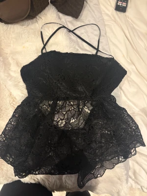 Svart spetsig linnetopp från Bik Bok - Säljer en svart linnetopp från Bik Bok med tunna axelband som korsas i ryggen. Toppen har ett genombrutet spetsmaterial med snygga detaljer och en lätt volang nedtill. Justerbara axelband för perfekt passform. 