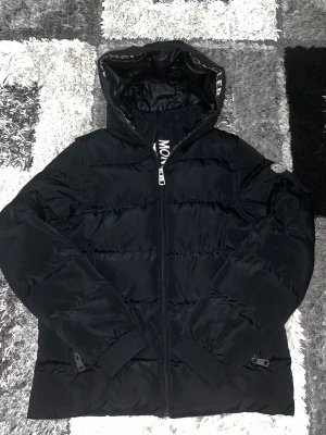 Svart Moncler pufferjacka med huva - Svart pufferjacka från Moncler med stor huva och logotyp på ärmen. Jackan har dragkedja framtill, ribbade muddar och Moncler-tryck på insidan av huvan. Fodret har ett coolt serietryck och jackan är tillverkad i blankt syntetmaterial med dunfyllning.