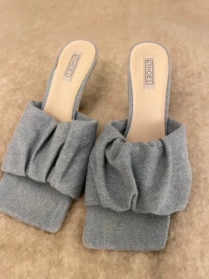 Blåa jeans klackskor NLY Shoes - Snygga klackskor från NLY Shoes i ljusblått tyg med breda band och jeansmatrial framtill. Skorna har fyrkantig tå och klack, vilket ger en trendig look. Perfekta för dig som gillar att sticka ut med detaljer och stilren design.