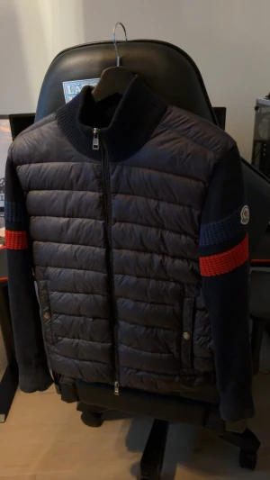 Moncler mörkblå cardigan med röd detalj - Säljer en mörkblå pufferjacka från Moncler med stickade ärmar och röda samt blå ribbade detaljer på ärmarna. Jackan har dragkedja framtill, hög krage och Moncler-logga på ena ärmen. Materialmix av polyester och stickad ull. Perfekt för dig som gillar street och exklusiva märken. Strl s men passar xs! Skadorna går att fixa hos skräddare. 