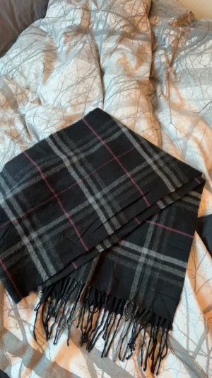 Rutig svart halsduk från Burberrys - Snygg halsduk från Burberrys i 100% lammull med klassiskt rutigt mönster i svart, grått och tunna röda linjer. Mjukt material och fransar i kanterna, perfekt att slänga över axlarna för en cool look. Aldrig använd! 