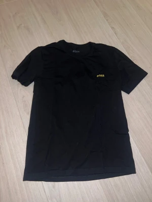 Svart t-shirt från BOSS - Säljer en svart t-shirt från BOSS med gul logotyp på bröstet.