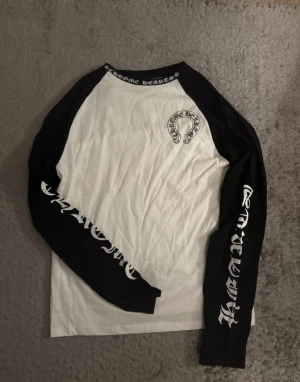 Chrome Hearts långärmad tröja svart/vit - Säljer en långärmad tröja från Chrome Hearts i svart och vitt med feta tryck på både ärmar och rygg. Tröjan har kontrastfärgade raglanärmar och coola logodetaljer runt halsen och på bröstet. Materialet är mjuk bomull, perfekt för dig som gillar streetwear.