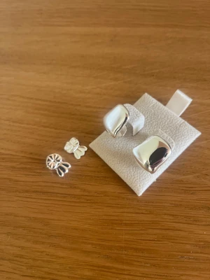 Snygga silvriga örhängen från Muli collection - Två par silvriga örhängen, ett par chunky och fyrkantiga med mjuka former och ett par små studs med blommig design. Båda är i blankt silverfärgat material och har en modern vibe. Perfekt för dig som gillar att mixa och matcha olika stilar.