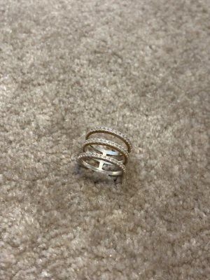 Trebandsring i silver med stenar - Säljer en snygg trebandsring i silverfärgat metall med glittrande små klara stenar på varje band. Ringen har en modern och öppen design som ger ett coolt statement på fingret. Perfekt för dig som gillar att sticka ut med dina smycken.
