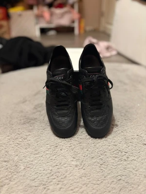 Svarta sneakers från Gucci med röd/grön detalj - Gucci sneakers i svart skinn med präglat GG-mönster och klassiska röd/gröna ränder på sidan. Rund tå, platt sula och snörning framtill. Snygg och stilren design med diskret logga på tungan och hälen. Perfekt för dig som gillar lyxiga streetwear-vibes.