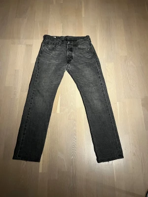 Levi's 501 svarta jeans straight fit - Svarta Levi's 501 jeans med klassisk straight fit och fem fickor. Jeansen har en knappgylf, tydlig Levi's-lapp bak och röd Levi's-tag på bakfickan. Materialet är slitstarkt denim i bomull och färgen är mörkgrå/svart med lätt tvättad look. 32/32