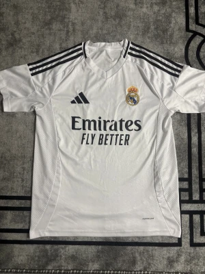Real Madrid vit matchtröja Adidas - Säljer en officiell Real Madrid fotbollströja från Adidas i vit färg med svarta detaljer och broderat klubbmärke. Tröjan har korta ärmar, V-ringad krage och tre svarta ränder på axlarna. Materialet är lätt och andas, perfekt för träning eller match.