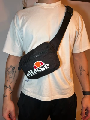Svart axelväska från Ellesse - Snygg svart axelväska från Ellesse med stort vitt och orange logotryck på framsidan. Väskan har en justerbar rem med plastspänne och en praktisk dragkedja upptill. Perfekt storlek för att bära med sig det viktigaste och passar till streetwear.