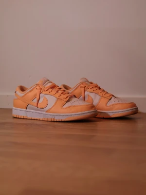 Nike Dunk Low orange och vita sneakers ”Peach White” - Säljer ett par Nike Dunk Low sneakers i en fräsch orange och vit färgkombination. Skorna har klassisk snörning, låg profil och en platt sula. Perfekta för dig som vill sticka ut med färg på fötterna. Ovandelen är i skinn och de har den ikoniska swoosh-loggan på sidan. ”Peach White”