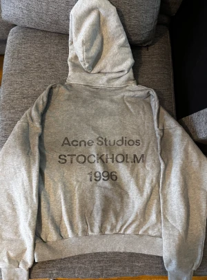 Grå hoodie Acne Studios Stockholm - Säljer en grå hoodie från Acne Studios med tryck 'Stockholm 1996' på ryggen. Tröjan har huva med slitna detaljer, ribbade muddar och är tillverkad i mjuk bomull med fleecefoder på insidan. Perfekt för dig som gillar clean streetwear med en twist.