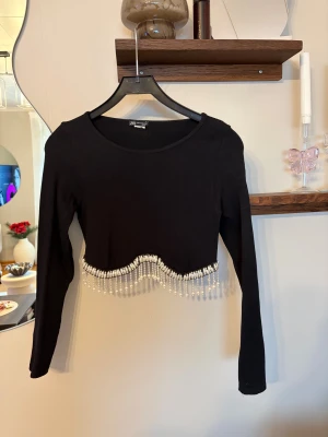 Svart croppad topp med pärldetaljer från Zara - Supersnygg svart croppad topp från Zara med långa ärmar och rund hals. Toppen är ribbad och har en vågig kant nedtill som är dekorerad med vita pärlor och pärlfransar som ger en extra cool touch. Perfekt för dig som vill sticka ut lite extra.