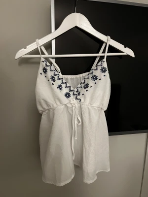 Vit broderad linnetopp med knyt - Säljer en söt vit linnetopp med tunna axelband och blå broderade blommor längs v-ringningen. Toppen har knyt i midjan och är tillverkad i ett lätt, luftigt material som passar perfekt för varma dagar. Stilren och fräsch look med fina detaljer.