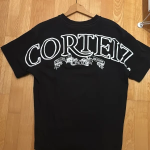 Svart Corteiz t-shirt - Svart t-shirt från Corteiz i storlek M med fet vit logga över ryggen och ett mindre emblem med tank och lagerkrans på bröstet. Klassisk rund hals och korta ärmar. Materialet är mjuk bomull som känns skönt mot huden. Perfekt för dig som gillar streetwear och vill sticka ut.