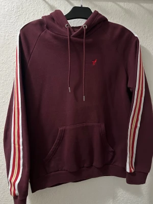 Vinröd hoodie från Pier One med ränder - Säljer en vinröd hoodie från Pier One med vita och röda ränder längs ärmarna. Tröjan har huva med snörning, magficka och ett litet rött broderat motiv på bröstet. Perfekt för dig som gillar sportig och avslappnad stil.