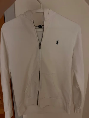 Vit hoodie från Polo Ralph Lauren - Vit hoodie från Polo Ralph Lauren med dragkedja och klassisk logga på bröstet. Tröjan har huva, två fickor framtill och är långärmad. Tillverkad i mjuk bomull som är skön att ha på sig. Perfekt för dig som gillar stilrena och enkla plagg. Nypris är ungefär 1300 kr. Skick 9,5/10 och mitt pris 750 kr. Pris kan diskuteras vid snabb affär. 