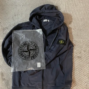 Mörkblå hoodie från Stone Island - Säljer en mörkblå hoodie från Stone Island med klassisk kompasspatch på ärmen. Tröjan har huva med snörning, dragkedja framtill och ribbade muddar. Materialet är mjuk bomull och den har en clean, normal passform. Perfekt för dig som gillar streetwear och snygga detaljer.