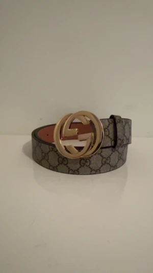 GG Supreme bälte med guldspänne - Säljer ett GG Supreme bälte med ikoniskt guldfärgat dubbel-G spänne. Bältet är beige och brunt med det klassiska GG-mönstret och har en bred, stilren form. Insidan är ljusbrun och materialet är skinn. Perfekt för dig som vill ha en lyxig detalj till outfiten.