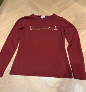 Vinröd långärmad tröja från Bondelid - Snygg vinröd långärmad tröja från Bondelid med guldigt tryck på bröstet. Tröjan har en klassisk rund hals och är gjord i mjuk bomull som är skön mot huden. Perfekt för dig som gillar stilrena och trendiga plagg med en touch av lyx.