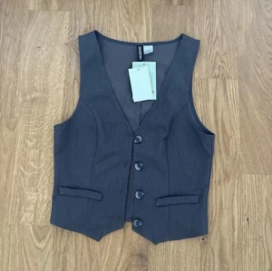 Mörkgrå kostymväst från Cropp - Snygg mörkgrå kostymväst från Cropp med klassisk v-ringning och fyra knappar framtill. Västen har två dekorativa fickor och är tillverkad i ett mjukt polyestermaterial som ger en stilren look. Perfekt att styla över en skjorta för en trendig outfit.