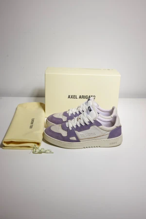 Axel Arigato dice lo - Säljer ett par snygga Axel Arigato sneakers i lila och vitt med låg profil. Skorna har en rund tå, platt sula och är tillverkade i skinn och mocka. De har vita snören och perforerade detaljer på tån. Perfekt för dig som gillar stilrena och trendiga sneakers. Extremt bra skick, ser helt nya ut!