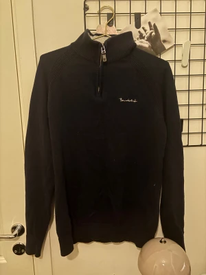 Svart stickad half zip-tröja Bondelid - Svart stickad tröja från Bondelid med half zip och hög krage. Diskret broderad logga på bröstet. Långärmad och ribbade detaljer vid axlar och muddar. Perfekt för dig som gillar stilren och enkel design.