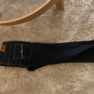 Mörkblå jeans från Nudie Jeans - Snygga mörkblå jeans från Nudie Jeans med klassisk femficksdesign och kontrastsömmar i orange. Modellen är Thin Finn och är storlek 32/32. Med dragkedja och knapp framtill. Bakfickorna har Nudie Jeans signaturbroderi i vitt. 