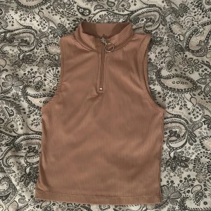 Beige ribbad ärmlös polotopp med zip - Säljer en beige ribbad polotopp med ärmlös design och en snygg silverfärgad dragkedja framtill med ringdetalj. Toppen är croppad och har en hög krage som ger en cool vibe. Perfekt att styla med jeans eller kjol för en trendig look.
