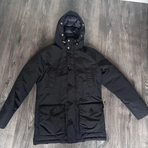 Svart parkajacka från J.Lindeberg - Svart parkajacka från J.Lindeberg med huva, stora fickor med lock och knappar samt snygg logga på ärmen. Jackan är vadderad och har både dragkedja och knappar framtill. Perfekt för kalla dagar när du vill hålla stilen.