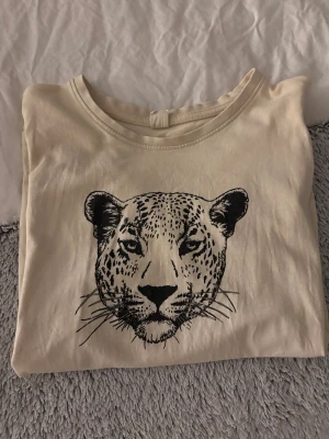 Beige t-shirt med leopardtryck H&M - Beige t-shirt från H&M med ett stort svart leopardhuvud tryckt på bröstet. Klassisk rund halsringning och korta ärmar. Skön och mjuk i bomull.